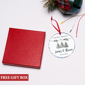 Op de afbeelding: Een heldere glazen kerstbal met een rood lint, met de tekst "Our First Christmas" en de namen "James & Sharon" met de datum "05.12.2024". Een rode geschenkdoos staat naast het ornament. De tekst "FREE GIFT BOX" staat onderaan.