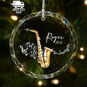 May include: A circular, faceted glass ornament featuring a gold saxophone illustration, white musical notes, and the text "Roger 2026" in a script font. The ornament includes a silver hanging cord attached to the top for display purposes.