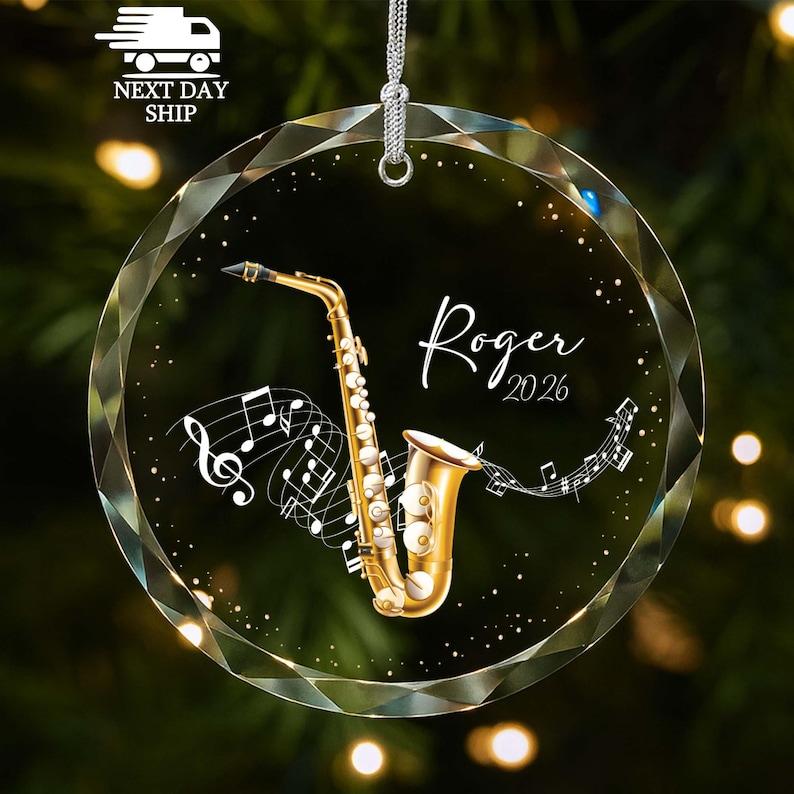 May include: A circular, faceted glass ornament featuring a gold saxophone illustration, white musical notes, and the text "Roger 2026" in a script font. The ornament includes a silver hanging cord attached to the top for display purposes.