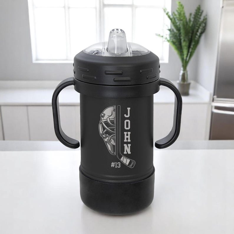 May include: Black insulated sippy cup with a clear, flip-top lid and two handles. The cup features a hockey-themed design with a goalie mask, hockey stick, the name "JOHN", and the number "13" etched on the side.