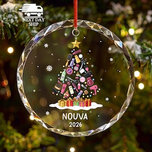 May include: A clear, circular, faceted glass ornament featuring a printed graphic of a Christmas tree composed of various hair salon tools, including scissors, brushes, and bottles. Below the tree are colorful gift boxes, the name NOUVA, and 2026.