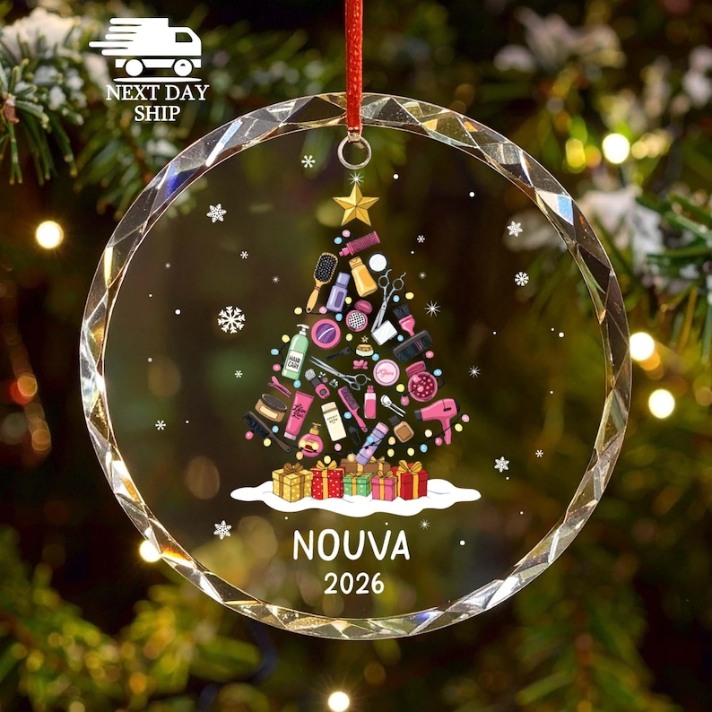 May include: A clear, circular, faceted glass ornament featuring a printed graphic of a Christmas tree composed of various hair salon tools, including scissors, brushes, and bottles. Below the tree are colorful gift boxes, the name NOUVA, and 2026.