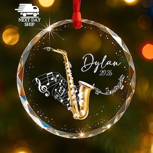 May include: A round, clear, faceted glass ornament featuring a gold saxophone illustration, white musical notes, and the text "Dylan 2026" in a script font. The ornament is suspended by a red ribbon against a blurred, warm-toned background.