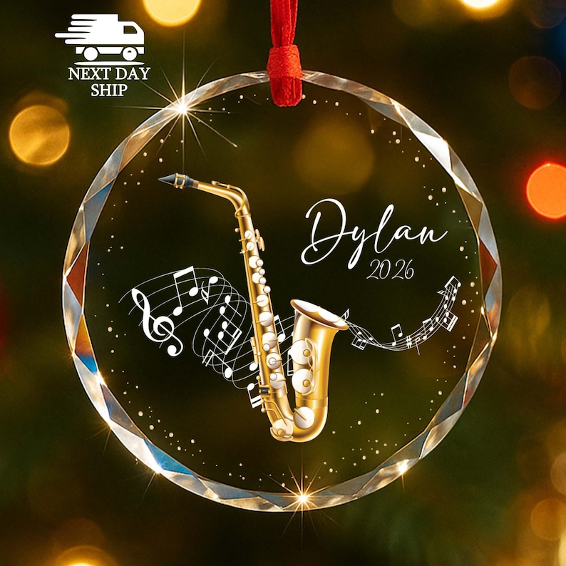 May include: A round, clear, faceted glass ornament featuring a gold saxophone illustration, white musical notes, and the text "Dylan 2026" in a script font. The ornament is suspended by a red ribbon against a blurred, warm-toned background.