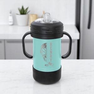 May include: A teal and black sippy cup with a hockey-themed design. The cup features a hockey helmet and stick graphic, the name "HENRY" vertically, and the number "#37". It has a black lid, handles, and base.