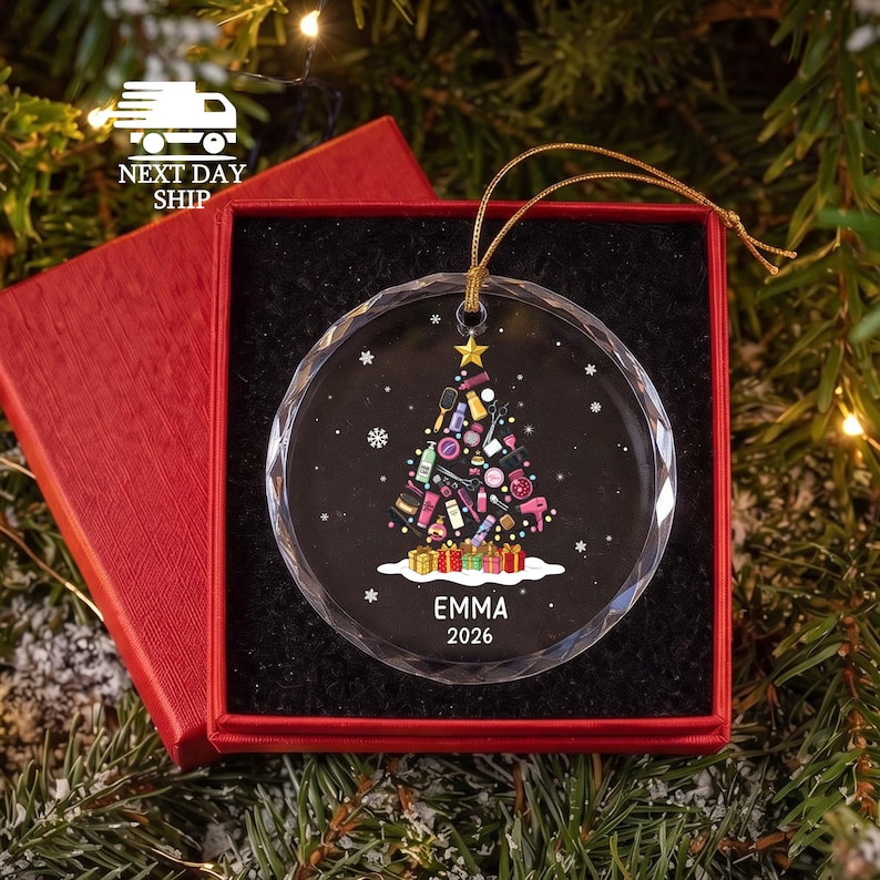 May include: A round, clear glass Christmas ornament with a faceted edge, featuring a printed graphic of a Christmas tree composed of various beauty and hair styling tools. The name EMMA and the year 2026 are printed in white below the tree.