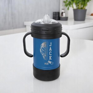 May include: A blue insulated tumbler with a black lid and handles. The tumbler features a hockey-themed design with the name "JACK" and the number "09" etched on the side. The tumbler has a clear, flip-top spout.