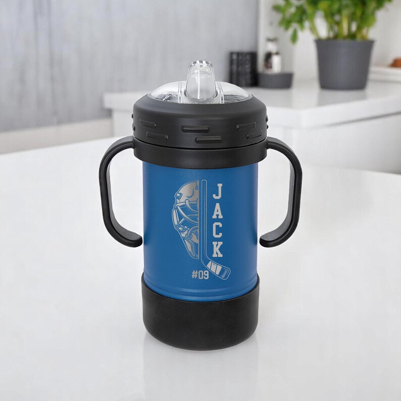May include: A blue insulated tumbler with a black lid and handles. The tumbler features a hockey-themed design with the name "JACK" and the number "09" etched on the side. The tumbler has a clear, flip-top spout.