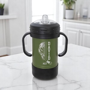 May include: An olive green sippy cup with black accents, featuring a hockey-themed design. The cup has a black lid and handles, with the name "DAVID" and the number "07" printed on the side. The design includes a hockey mask and a hockey stick.