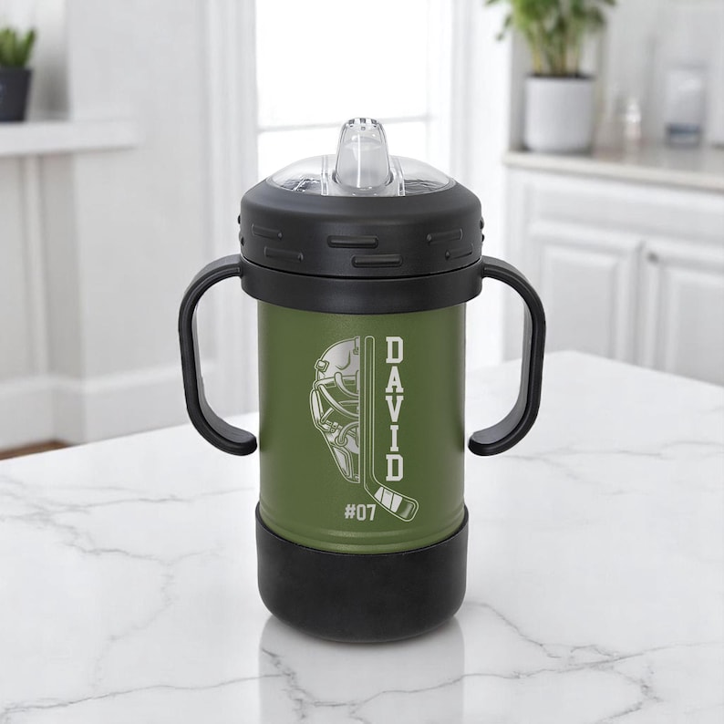 May include: An olive green sippy cup with black accents, featuring a hockey-themed design. The cup has a black lid and handles, with the name "DAVID" and the number "07" printed on the side. The design includes a hockey mask and a hockey stick.
