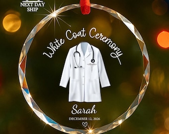 Puede incluir: Un adorno de cristal circular facetado con una bata blanca, un estetoscopio negro y un símbolo de caduceo médico. El texto dice White Coat Ceremony, Sarah y December 12, 2026, con un pequeño icono de corazón, suspendido por una cinta roja.
