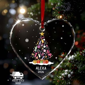 May include: A heart-shaped clear acrylic ornament featuring a Christmas tree graphic composed of various hair styling tools and beauty products. The design includes a gold star topper, colorful gift boxes at the base, and the text ALEXA 2026.