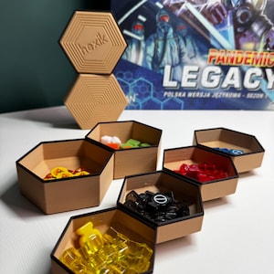 Op de afbeelding: Een bordspelopstelling met zeshoekige houten bakjes gevuld met spelstukken in verschillende kleuren. De spelstukken zijn geel, rood, groen en zwart. De speldoos, getiteld "Pandemic Legacy", is zichtbaar op de achtergrond.