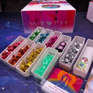 Può includere: Una scatola di gioco da tavolo con l'etichetta WROTH si trova dietro un vassoio di plastica contenente vari componenti di gioco. Il vassoio contiene piccoli gettoni di plastica colorati in rosso, arancione, giallo, verde, verde acqua, blu, magenta e argento.