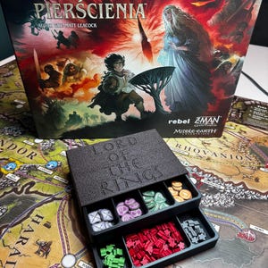 Puede incluir: Una caja de juego de mesa con el título "Lord of the Rings" en inglés y polaco. La caja presenta ilustraciones de personajes y escenas del mundo de fantasía. Un organizador negro con piezas de juego se encuentra sobre un mapa de la Tierra Media.