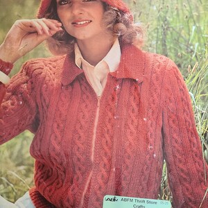 Può includere: Una donna indossa un cappello lavorato a maglia color ruggine e un maglione con zip abbinato. Il maglione presenta un motivo a trecce e un colletto. L'immagine è una fotografia d'epoca, probabilmente da un modello artigianale o da una rivista.