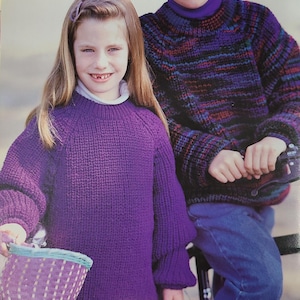 May include: Two children wearing knitted jumpers. The girl is wearing a purple jumper and the boy is wearing a multi-coloured jumper with a striped pattern. They are both smiling and looking at the camera.