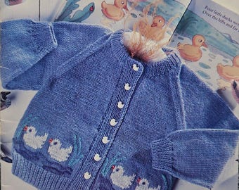 May include: A blue knitted cardigan with a white button closure and a duck design on the front. The cardigan is made with Patons Astra yarn. The image also shows a book with the title "Happy Days" and a price tag of $2.95.