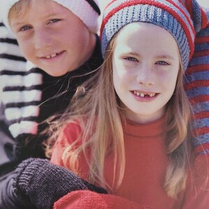 May include: Two children wearing striped knitted hats. The girl is wearing a red jumper and a pair of black and red knitted mittens. The boy is wearing a black and white striped scarf.