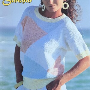 May include: A woman in a white, blue, and peach-coloured knit sweater with short sleeves. The sweater has a geometric design and is paired with white shorts. The image also features the text "Beehives", "Patons Cotton Sahara".