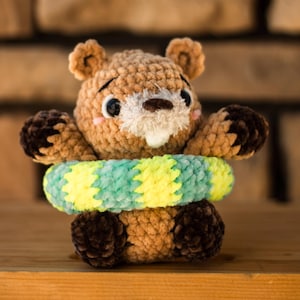 May include: A handmade crochet groundhog toy in shades of brown, with a white muzzle and a green and yellow striped ring. The plush toy has dark brown paws and ears, and is sitting on a wooden surface.
