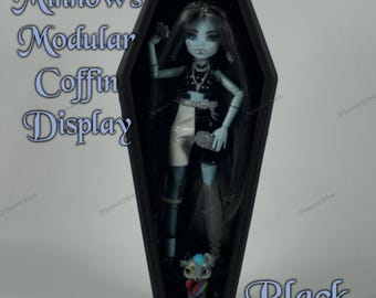 May include: A black coffin-shaped display case with a doll inside. The doll has blue skin, long black hair, and is wearing a black and white outfit. The text "Minnow's Modular Coffin Display" is visible on the left side, and "Black" is on the bottom right.