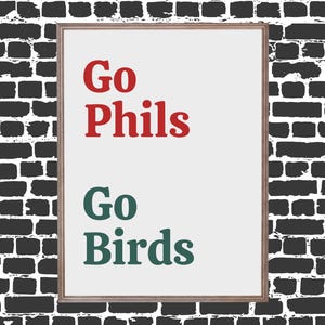 May include: A framed print with the words "Go Phils" in red and "Go Birds" in green. The print has a white background and a wooden frame, set against a black brick wall.
