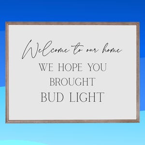 Può includere: Un cartello incorniciato con la scritta "Welcome to our home. We hope you brought Bud Light" in nero su sfondo bianco. La cornice è di colore marrone chiaro e lo sfondo è sfumato blu.