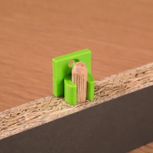 Puede incluir: Un conector de muebles de plástico verde con una clavija de madera insertada, fijado a un tablero de partículas. El conector es rectangular con un agujero circular y dos brazos curvos. El tablero tiene un borde oscuro y una superficie texturizada marrón claro.