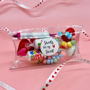 May include: A clear plastic pillow box filled with assorted sweets, including heart-shaped sweets, a candy cane, and a roll of colourful candy. The box has a sticker that says "Sweets for my Sweet" with a red heart.