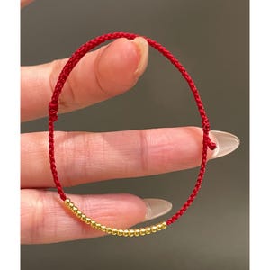 May include: A red string bracelet with a row of small, gold-colored beads. The bracelet is held by a hand, showcasing the woven red cord and the metallic beads. The design is simple and elegant.
