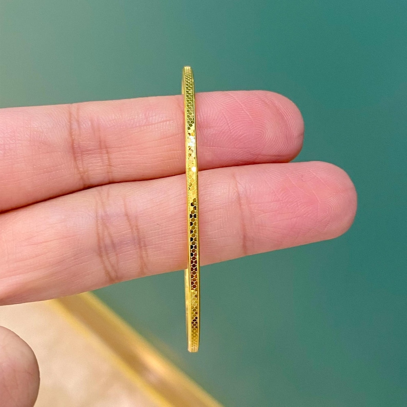 May include: A thin, gold-coloured bracelet with a textured surface. The bracelet is held between two fingers, showcasing its delicate design and intricate details. The bracelet appears to be made of metal and has a repeating pattern along its edges.
