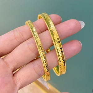 May include: Two gold bangles with a textured, geometric pattern. The bangles are held in a hand, showcasing their bright, reflective surface. The design features a series of small, connected shapes, creating a shimmering effect. The jewelry is a warm, golden colour.