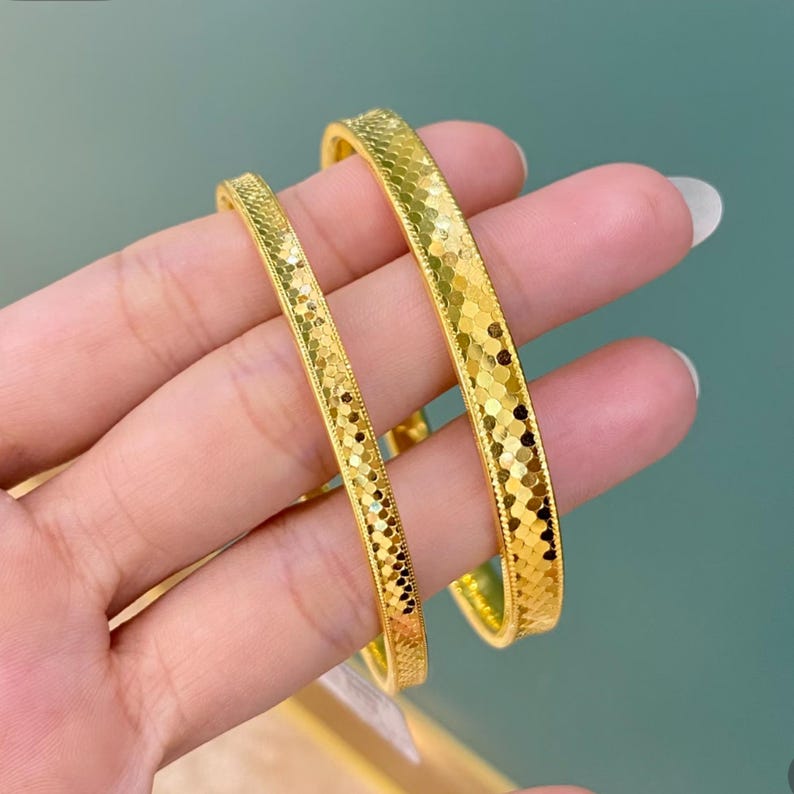 May include: Two gold bangles with a textured, geometric pattern. The bangles are held in a hand, showcasing their bright, reflective surface. The design features a series of small, connected shapes, creating a shimmering effect. The jewelry is a warm, golden colour.