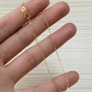 Könnte beinhalten: Eine zarte goldene Halskette mit einem Karabinerverschluss und kleinen, kreisförmigen Gliedern. Die Kette wird von einer Hand gehalten, die ihre feinen Details und ihre warme, goldene Farbe zeigt. Die Halskette ist für den täglichen Gebrauch konzipiert.