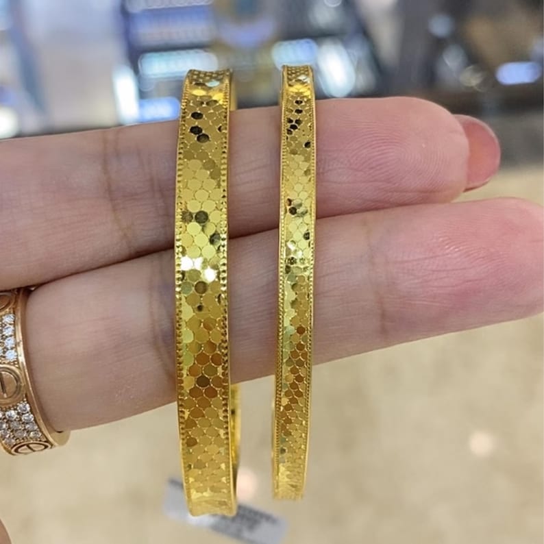 May include: Two gold bangles with a textured, honeycomb-like pattern. The bangles are held in a hand, showcasing their bright, reflective surface. The jewelry is a warm, yellow gold colour, and the design is consistent across both pieces.