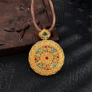May include: A gold pendant necklace with an intricate circular design. The pendant features red, blue, and green gemstones. The necklace has a brown woven cord.