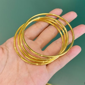 K&ouml;nnte beinhalten: Nahaufnahme einer Hand, die sechs goldfarbene Armreifen h&auml;lt. Die Armreifen sind d&uuml;nn, rund und gl&auml;nzend und reflektieren das Licht. Der Hintergrund ist einfarbig gr&uuml;n.