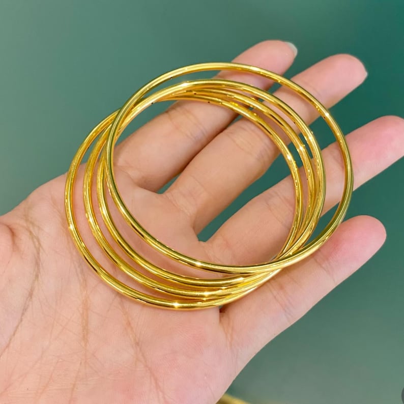 K&ouml;nnte beinhalten: Nahaufnahme einer Hand, die sechs goldfarbene Armreifen h&auml;lt. Die Armreifen sind d&uuml;nn, rund und gl&auml;nzend und reflektieren das Licht. Der Hintergrund ist einfarbig gr&uuml;n.