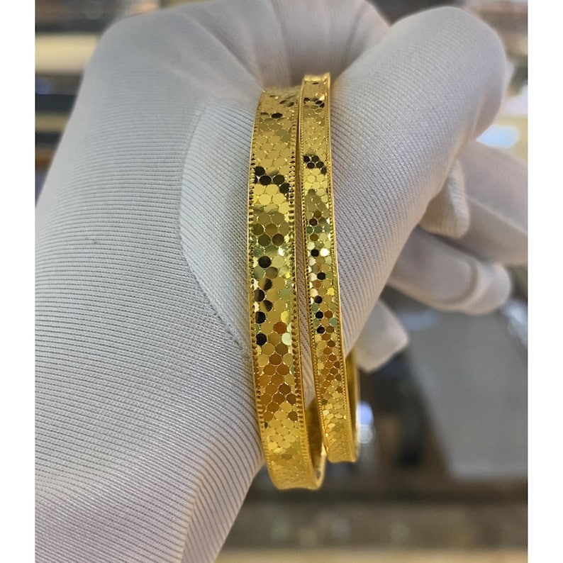 May include: Two gold bangles with a textured, dotted pattern. The bangles are held by a gloved hand. The gold bangles have a shiny finish and a repeating pattern of small, irregular shapes. The bangles are a bright, warm gold colour.