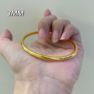 K&ouml;nnte beinhalten: Ein goldfarbenes Armreif-Armband mit einer Dicke von 3 mm. Das glatte, abgerundete Armband wird in einer Hand vor einem neutralen Hintergrund gehalten. Das Armband hat eine gl&auml;nzende, reflektierende Oberfl&auml;che.