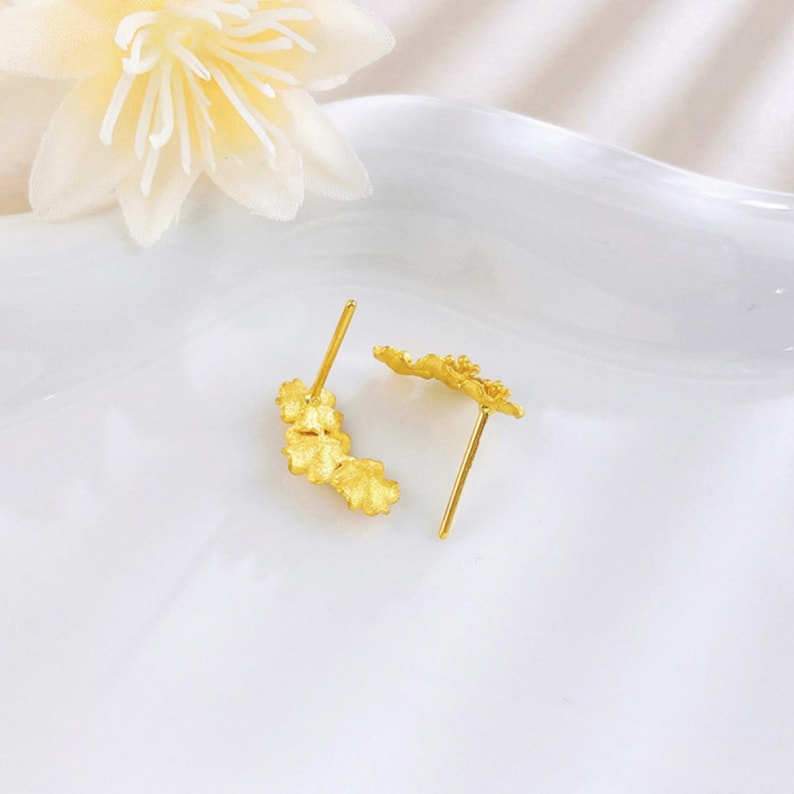 Puede incluir: Un par de pendientes de bot&oacute;n dorados con un dise&ntilde;o floral texturizado de varios p&eacute;talos. Cada pendiente tiene un poste fino y un acabado de superficie mate y granulado que capta la luz, presentado junto a una flor de color amarillo p&aacute;lido.