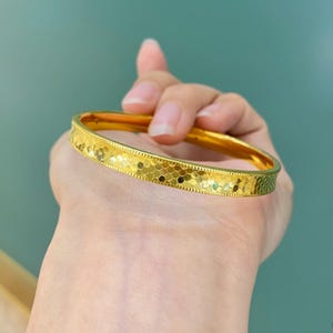 May include: A gold-coloured bangle bracelet with a textured, patterned surface. The bracelet is round and appears to be made of metal. The design features a repeating pattern of small, irregular shapes. The background is a solid teal colour.