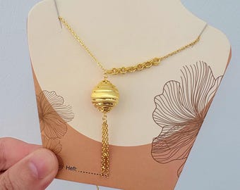 May include: A gold-colored necklace featuring a spherical pendant with horizontal ridges and a dangling tassel. The necklace is displayed on a beige card with floral designs and the word "Heft:" printed on it.