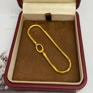 May include: A gold-colored bracelet with a snake chain design, resting inside a red jewelry box with a brown interior. The bracelet features a circular clasp and is presented in a well-lit setting.