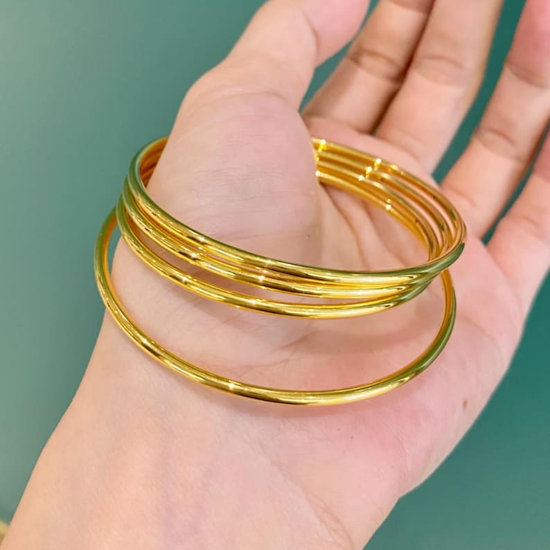 K&ouml;nnte beinhalten: Ein Satz goldfarbener Armreifen, &uuml;bereinander gestapelt, werden in einer offenen Hand gezeigt. Die Armreifen haben eine glatte, polierte Oberfl&auml;che, die das Licht reflektiert. Der Hintergrund ist einfarbig t&uuml;rkis.