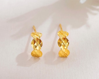 May include: A pair of gold-colored stud earrings. Each earring features a heart-shaped top, a central oval section with a textured design, and a heart-shaped bottom. The earrings are set against a white background.