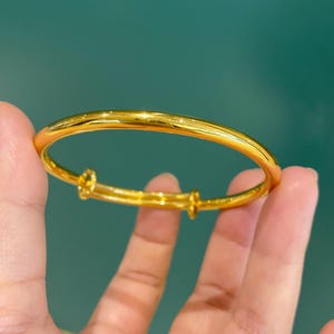 May include: A gold adjustable bangle bracelet. The bracelet is held in a hand against a teal background. The bracelet has a simple, elegant design, with a smooth, polished surface. The adjustable feature allows for a comfortable fit.