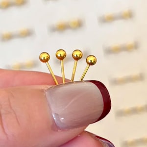 May include: Four gold-colored nail art studs, each with a small ball on top, are arranged in a row. The studs are held by a person's fingernail, which is painted with a light pink and burgundy nail polish.