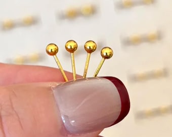 Puede incluir: Cuatro tachuelas doradas para decoración de uñas, cada una con una pequeña bola en la parte superior, dispuestas en fila. Las tachuelas son sostenidas por una uña, pintada con esmalte de uñas rosa claro y burdeos.